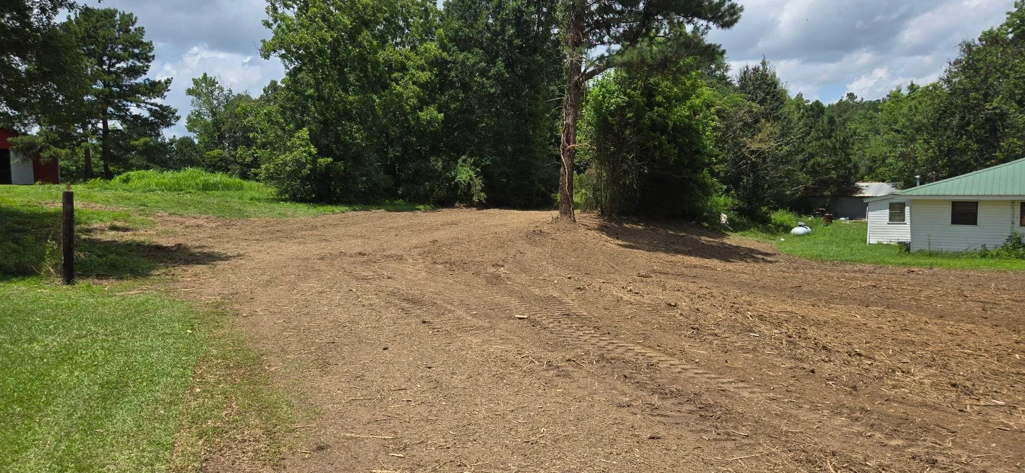 Cleared dirt lot with removed vegetation, mature trees, and residential structures visible