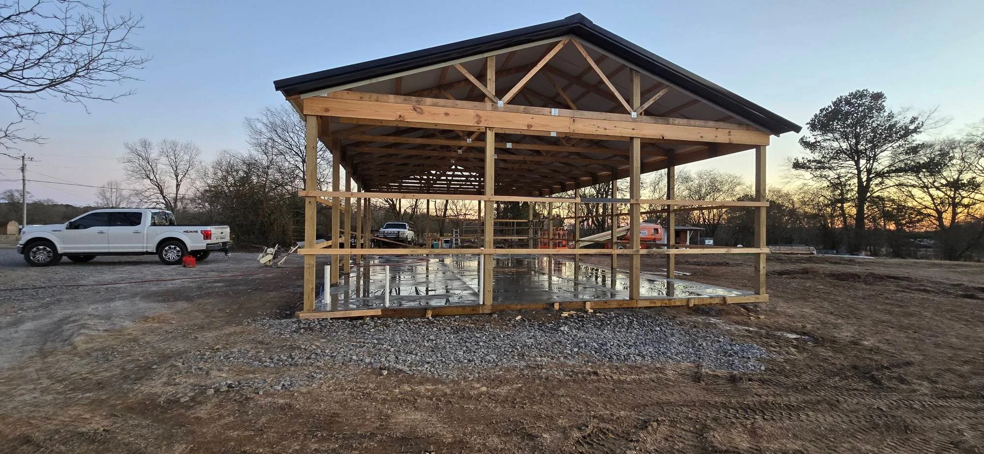 Timber frame pavilion with exposed beams, gabled roof, and concrete foundation on cleared land